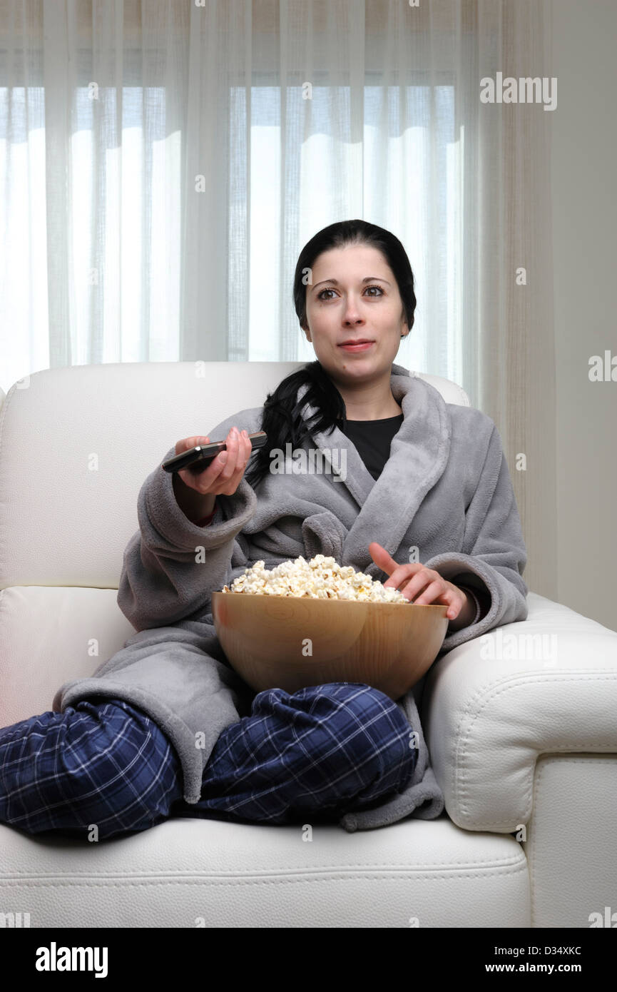 Young woman watching TV at home sitting on a white sofa and holding remote control Stock Photo