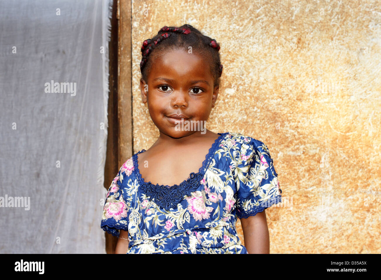 Portrait of a cute and sweet little black African girl, smiling but looking a bit shy, posing in front of her house. Stock Photo