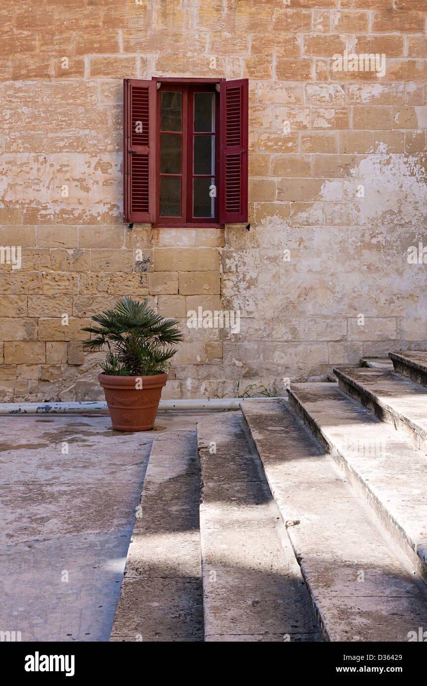 A shaft of light is cast on a set of stone steps, in the background is a lone window in a stone wall and small pot plant. Stock Photo