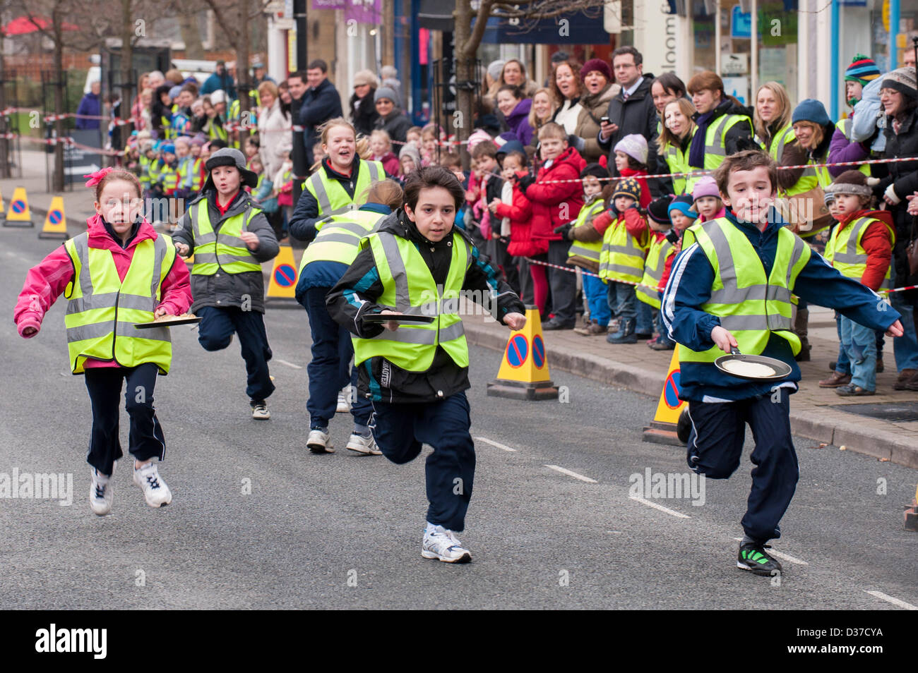 Crowd watches young competitors (children) taking part, running & flipping pancakes in traditional, Pancake Race - The Grove, Ilkley, West Yorkshire, UK. Stock Photo