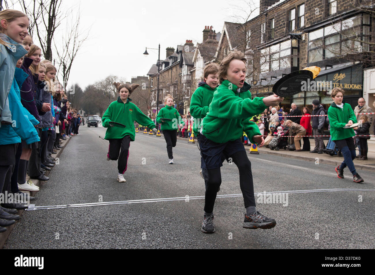 Crowd watches young competitors (children) taking part, running & flipping pancakes in traditional, Pancake Race - The Grove, Ilkley, West Yorkshire, UK. Stock Photo