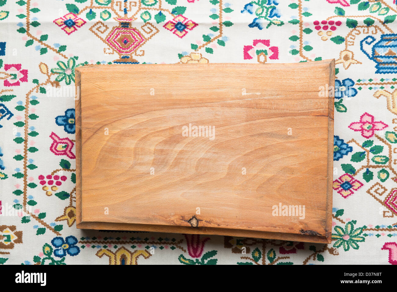 Natural wood chopping board on Danish folk fabric lit from the top of the frame with a drop shadow beneath the wooden block. Stock Photo