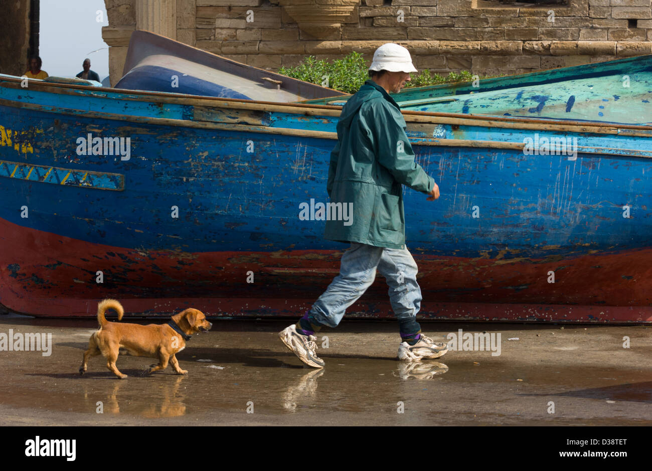Fisherman being followed by a dog, walking past a traditional blue fishing boat on the side of Essaouira Harbour, Essaouira, Morocco Stock Photo