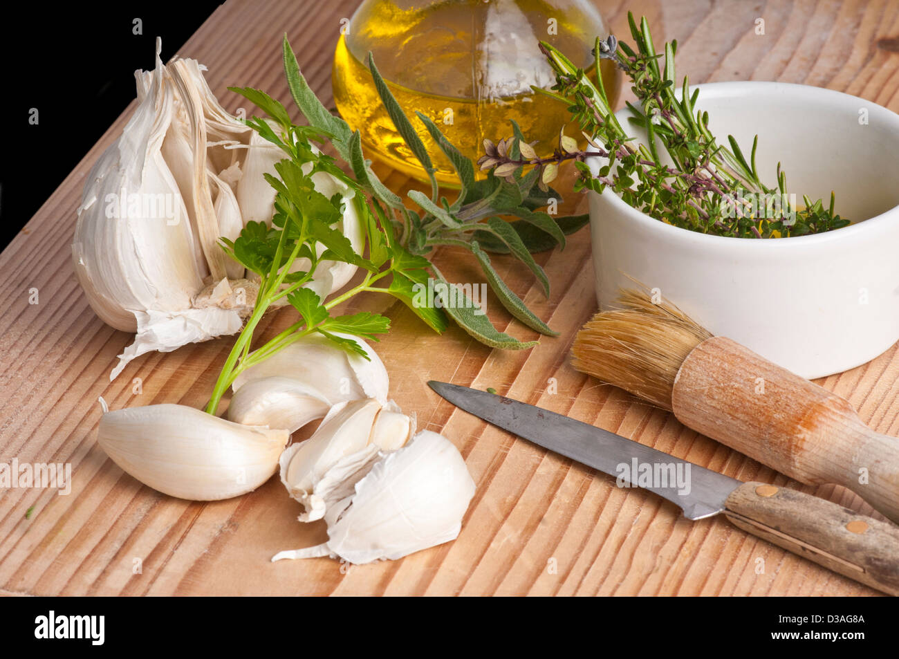 Garlic, parsley, sage, rosemary and thyme with extra virgen olive oil in a jug, a brush and knife. on an antique chopping board. Stock Photo