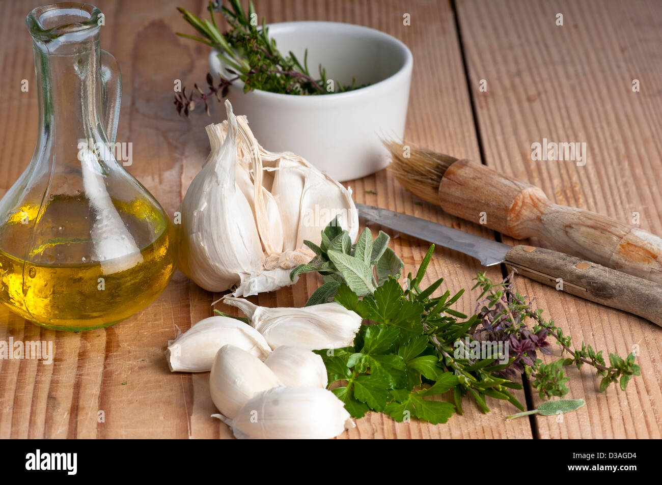 Garlic, parsley, sage, rosemary and thyme with extra virgin olive oil in a jug, a brush and knife. on an antique chopping board. Stock Photo