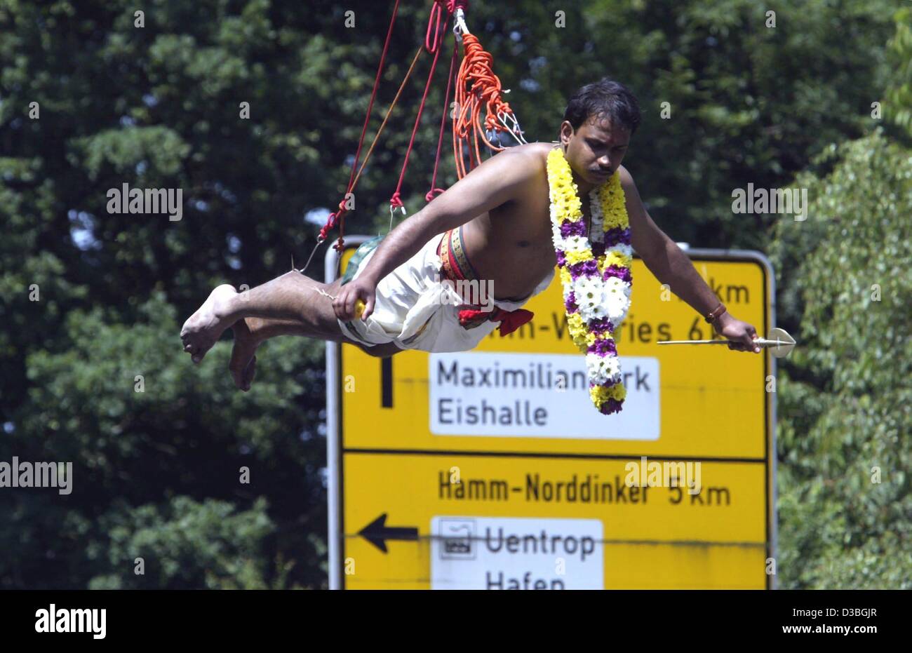 (dpa) - A believer of the Hindu faith hangs, attached to hooks which a pierced through the skin on his legs and back, on ropes and hovers in front of a yellow road sign in Hamm, Germany, 15 June 2003. He is part of a Hindu temple congregation which celebrates the goddess Sri Kamadchi Ampal (the godd Stock Photo