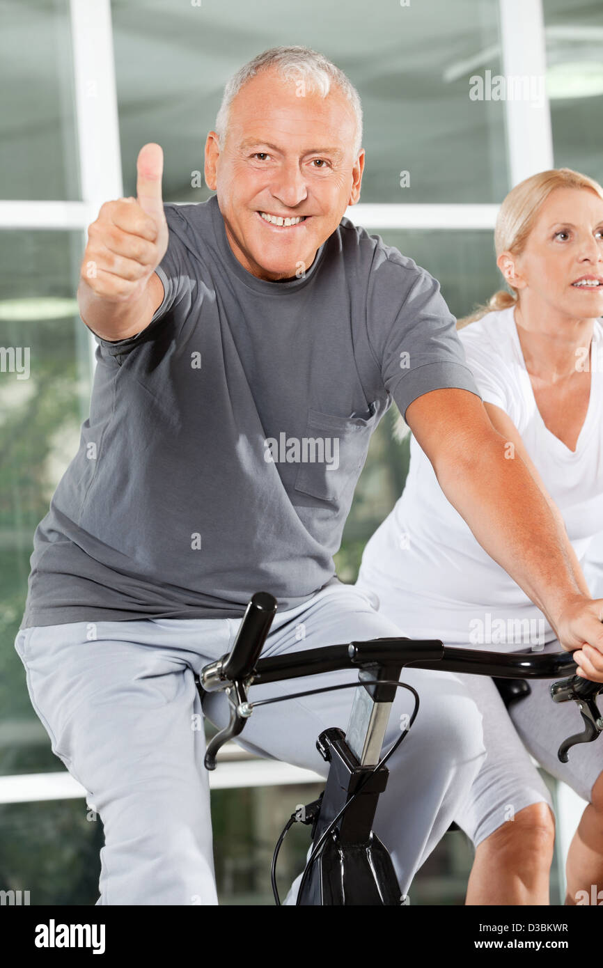 Happy senior man holding thumbs up on spinning bike in fitness center ...