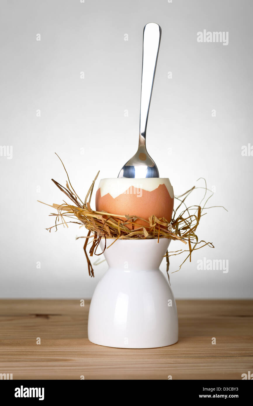 Easter egg in hay nest on white stand. Composition with egg ready to eat and spoon on wooden table background. Front view Stock Photo