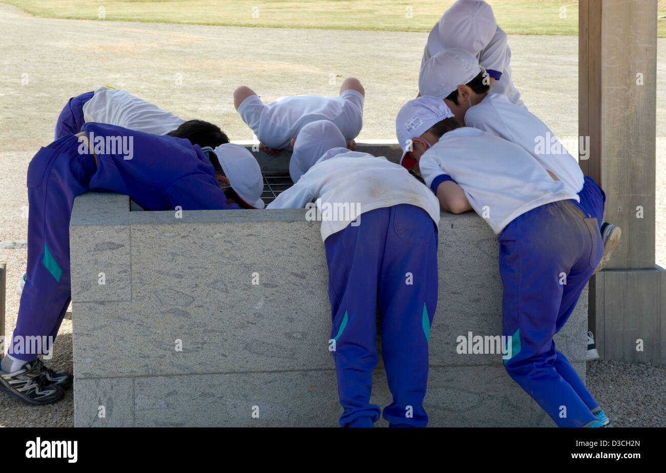 Group of curious, young Japanese school boys in sports uniforms bend over edge of a well as they explore the world around them. Stock Photo