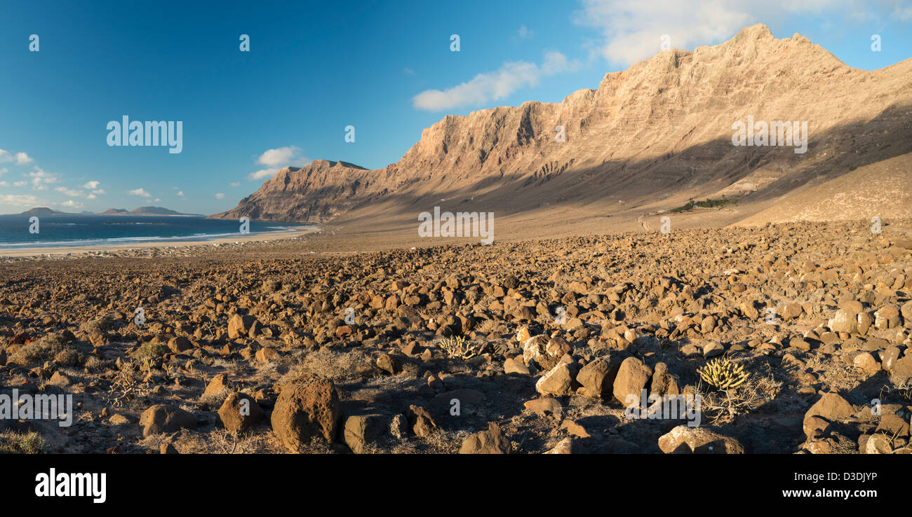 Malpais (rough ground or badland with dry scrub) in front of Famara cliff and beach at Caleta de Famara, Lanzarote Stock Photo
