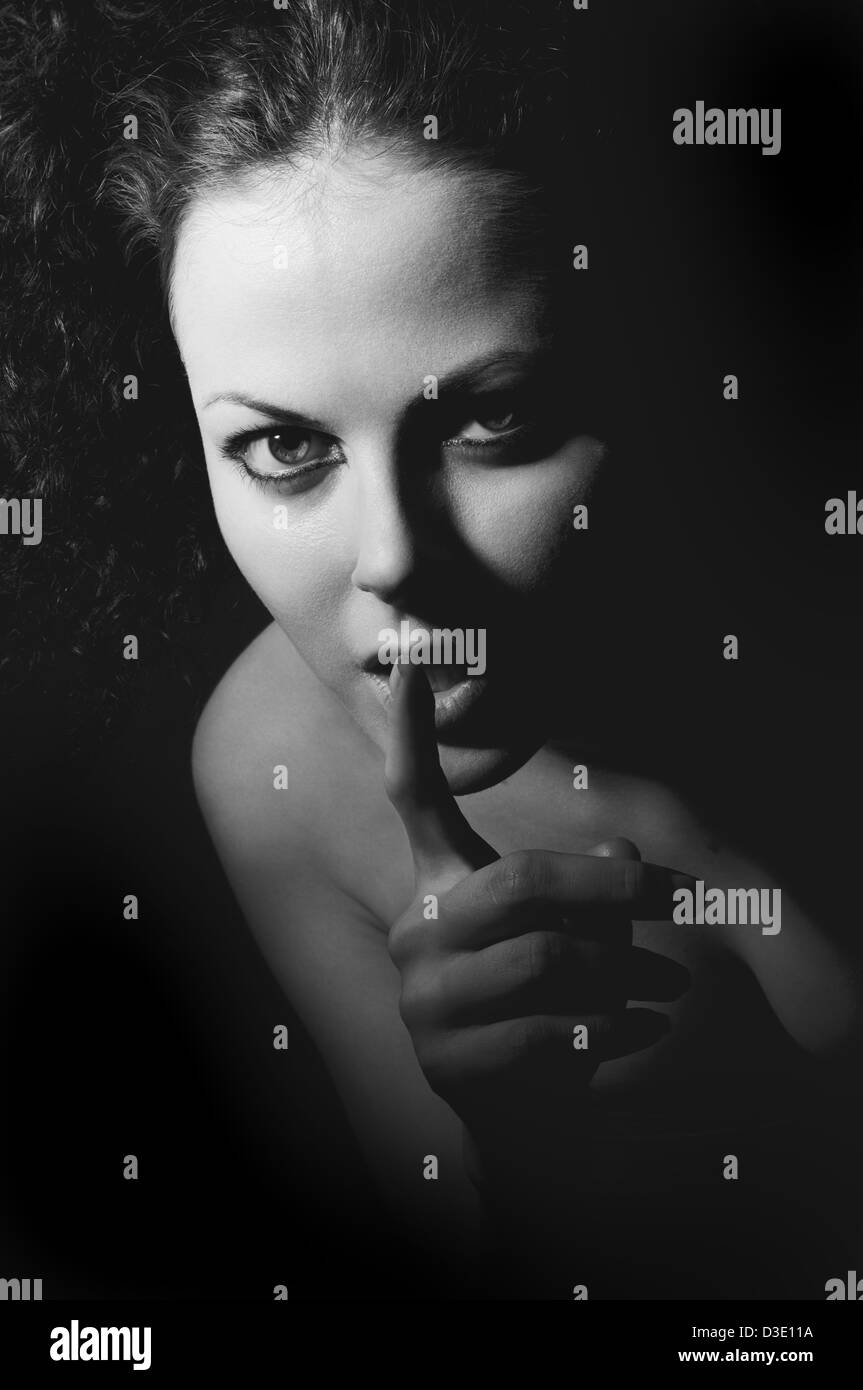 Young woman gesturing for being quiet, shows silence sign on dark background, black-white image Stock Photo