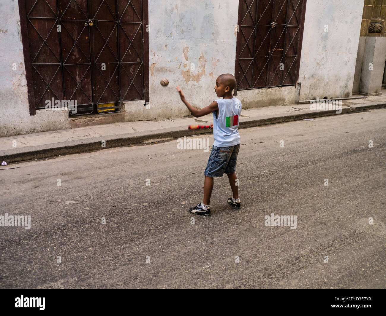 A young Cuban boy throws a baseball in the air as he starts his swing to hit it as he plays in the in the middle of the street. Stock Photo