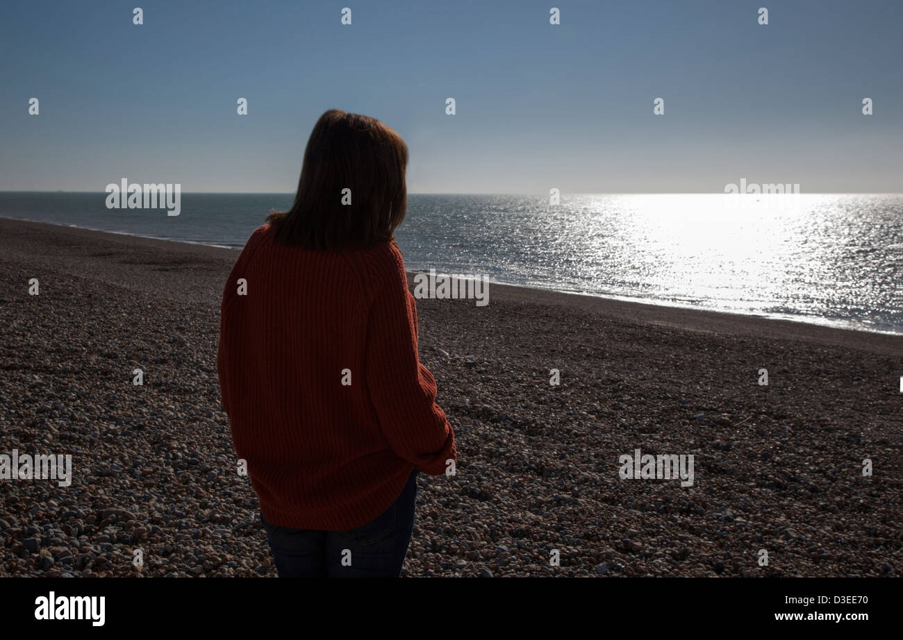 Back to camera silhouette of a woman wearing a chunky men's jumper looking out to sea, watching the sun glimmer on the water. Stock Photo