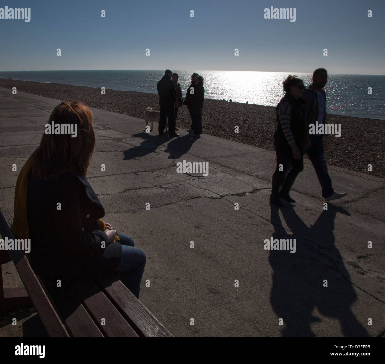 Silhouetted over the shoulder of a single woman sitting on a seat, watching people walking past and chatting to each other. Stock Photo
