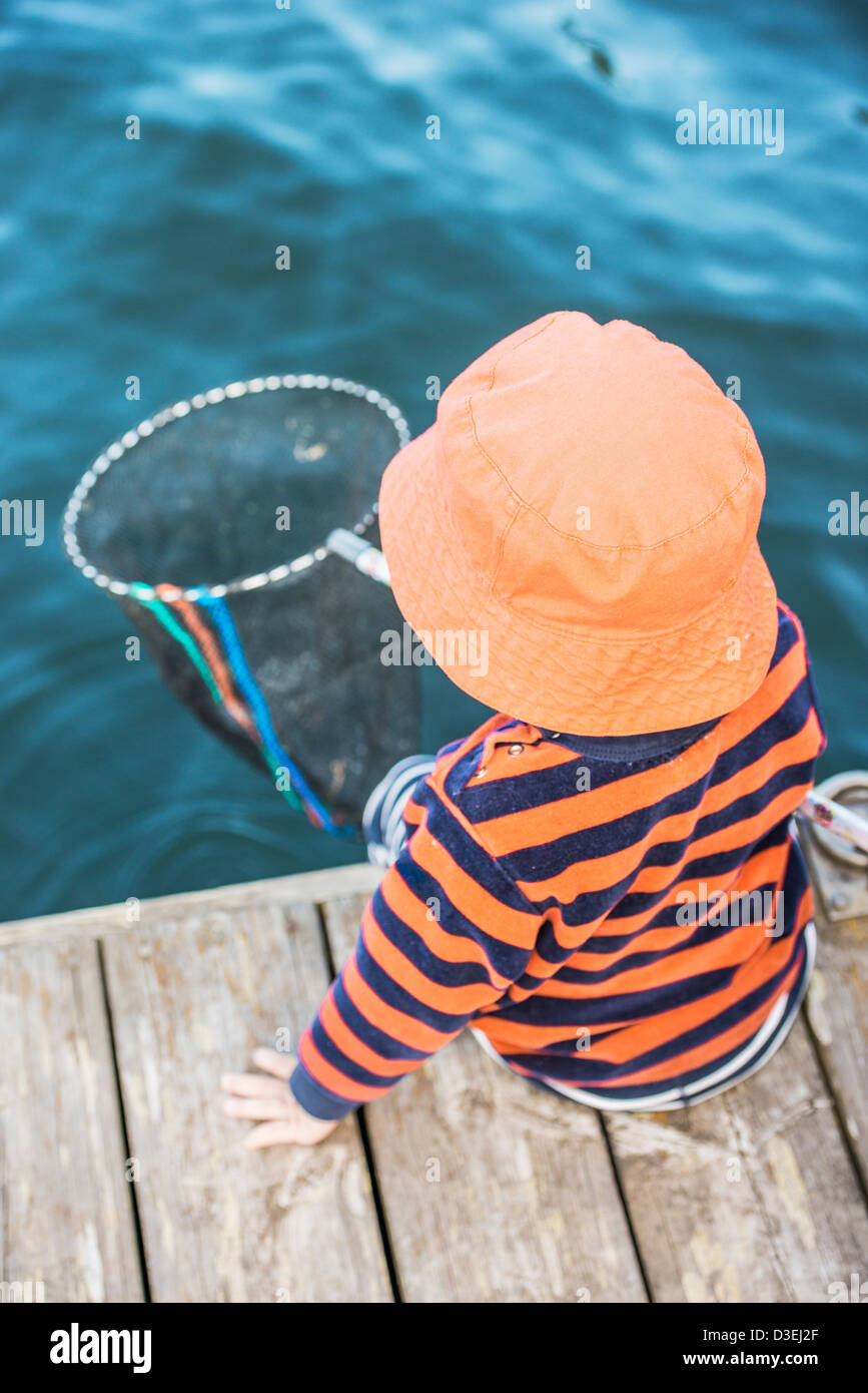 Young child sitting on dock by the sea watching the water with fishing net in hand Stock Photo
