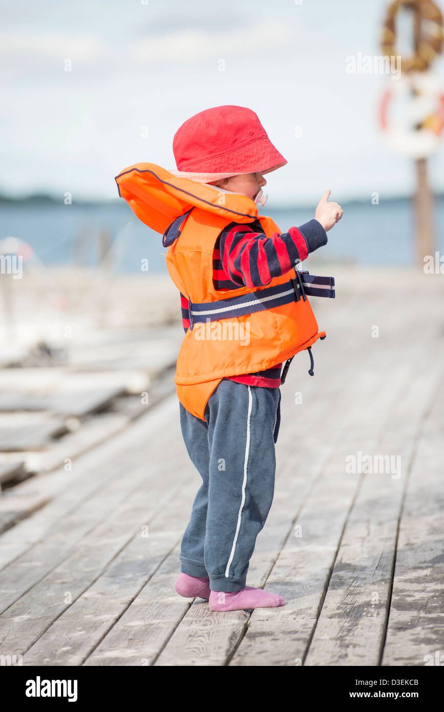 Young child wearing orange life jacket and red hat standing on wooden jetty by the sea Stock Photo