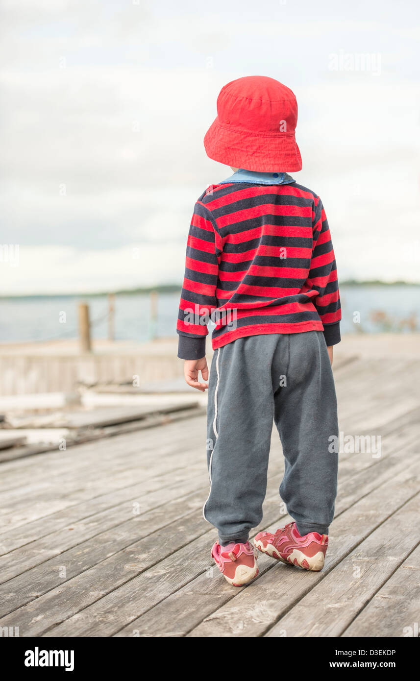 Young child wearing casual clothes and a red hat watching the sea from a wooden jetty Stock Photo