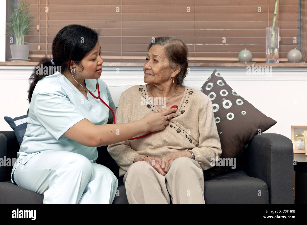 Home care provider keeps senior citizen patient company, checking her heartbeat with a stethoscope Stock Photo