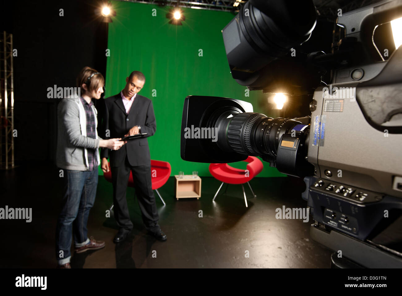 Foreground Television camera with floor manager and presenter out-of-focus in the background. Stock Photo