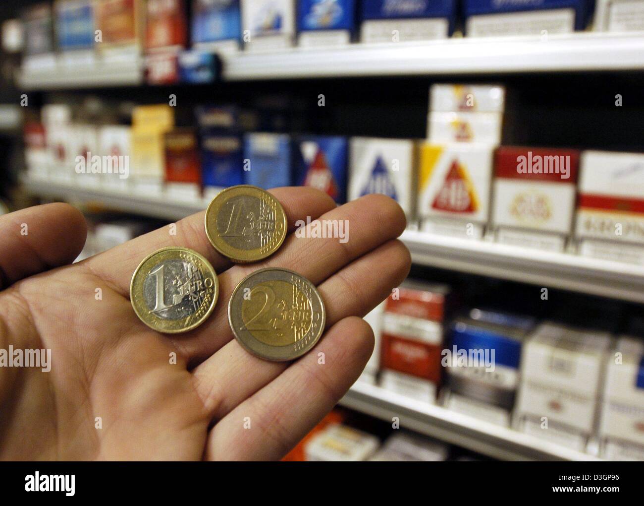 (dpa) - A hand holds up euro coins in front of a shelf of cigarette packs in a tobacco shop in Regensburg, Germany, 4 March 2004. The price of one pack with 19 cigarettes was raised 40 cents on 1 March 2004 in Germany. One pack now costs between 3.40 to 3.60 euros. The higher price is due to an increase of 1.2 cents of tax on each cigarette. The tax will contribute to the budget of Stock Photo