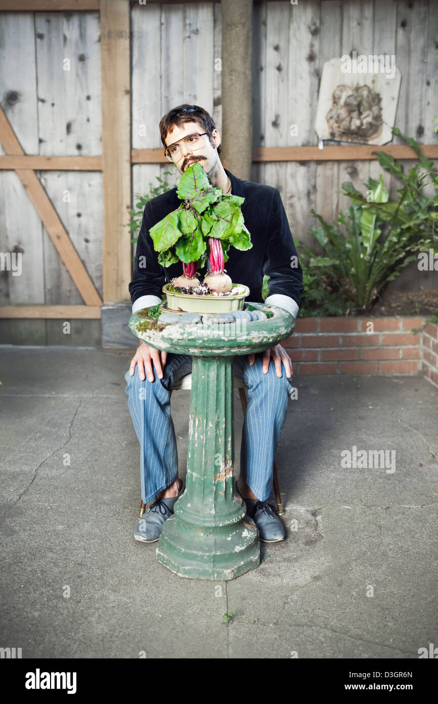 Hipsters and Beatniks - A young hipster man with mustache and glasses eating still planted beets on a birdbath in his own garden Stock Photo