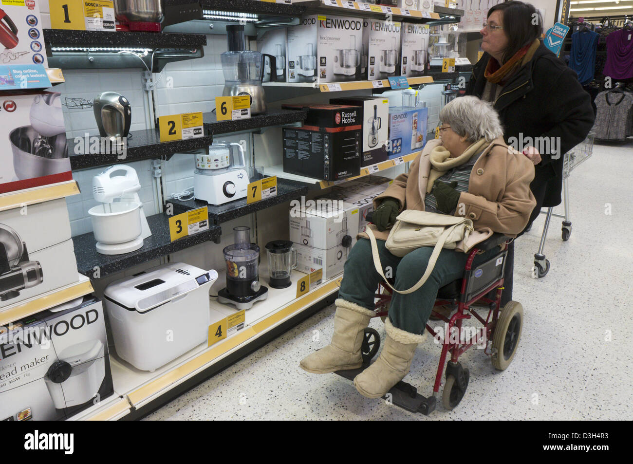 An elderly lady in a wheelchair and her carer or assistant look at kitchenware electrical products in a supermarket. Stock Photo