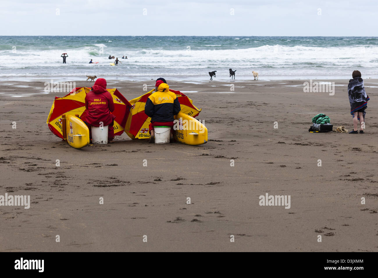 RNLI lifeguards and a small boy shelter from the cold wind on Black Rock Beach, Widemouth Bay, nr Bude, Cornwall, UK Stock Photo