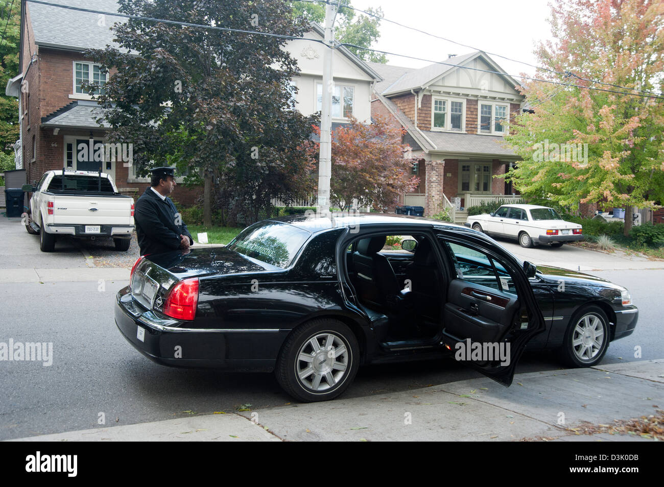 A limousine driver and his car wait at the curb of a residential neighborhood with door to car open waiting for passenger Stock Photo