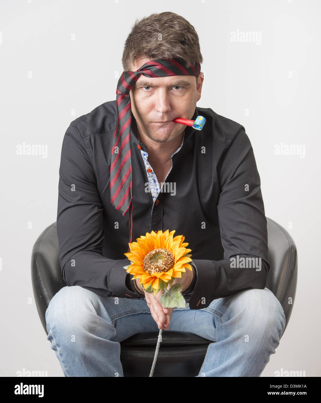 Bored businessman looking depressed at party. Sitting in armchair holding a plastic flower and wearing the tie on his forehead. Stock Photo