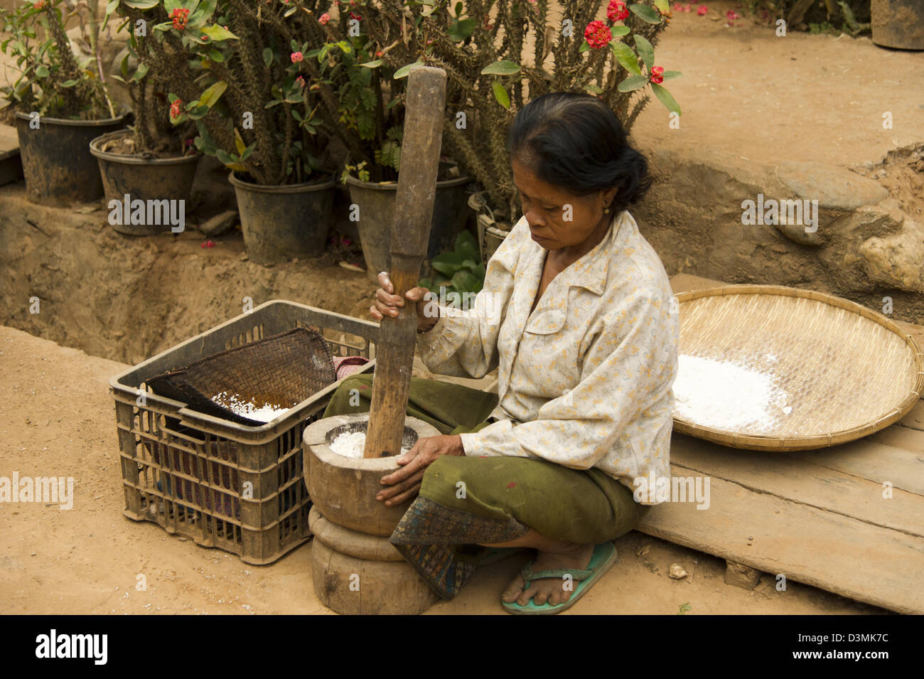A woman sits on steps preparing rice flour using a large mortar and