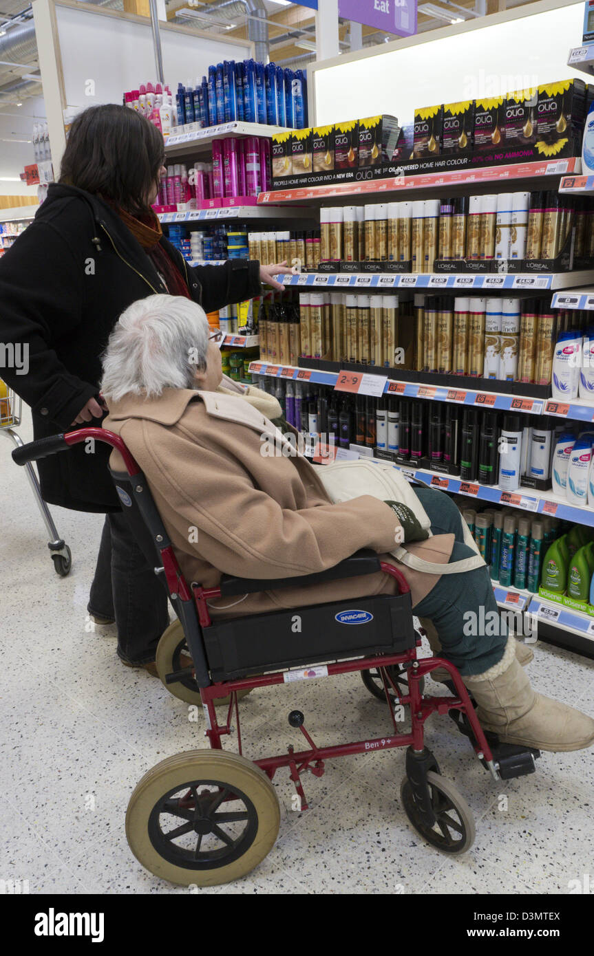 An elderly lady in a wheelchair and her carer or assistant look at hair products and shampoo in a supermarket. Stock Photo
