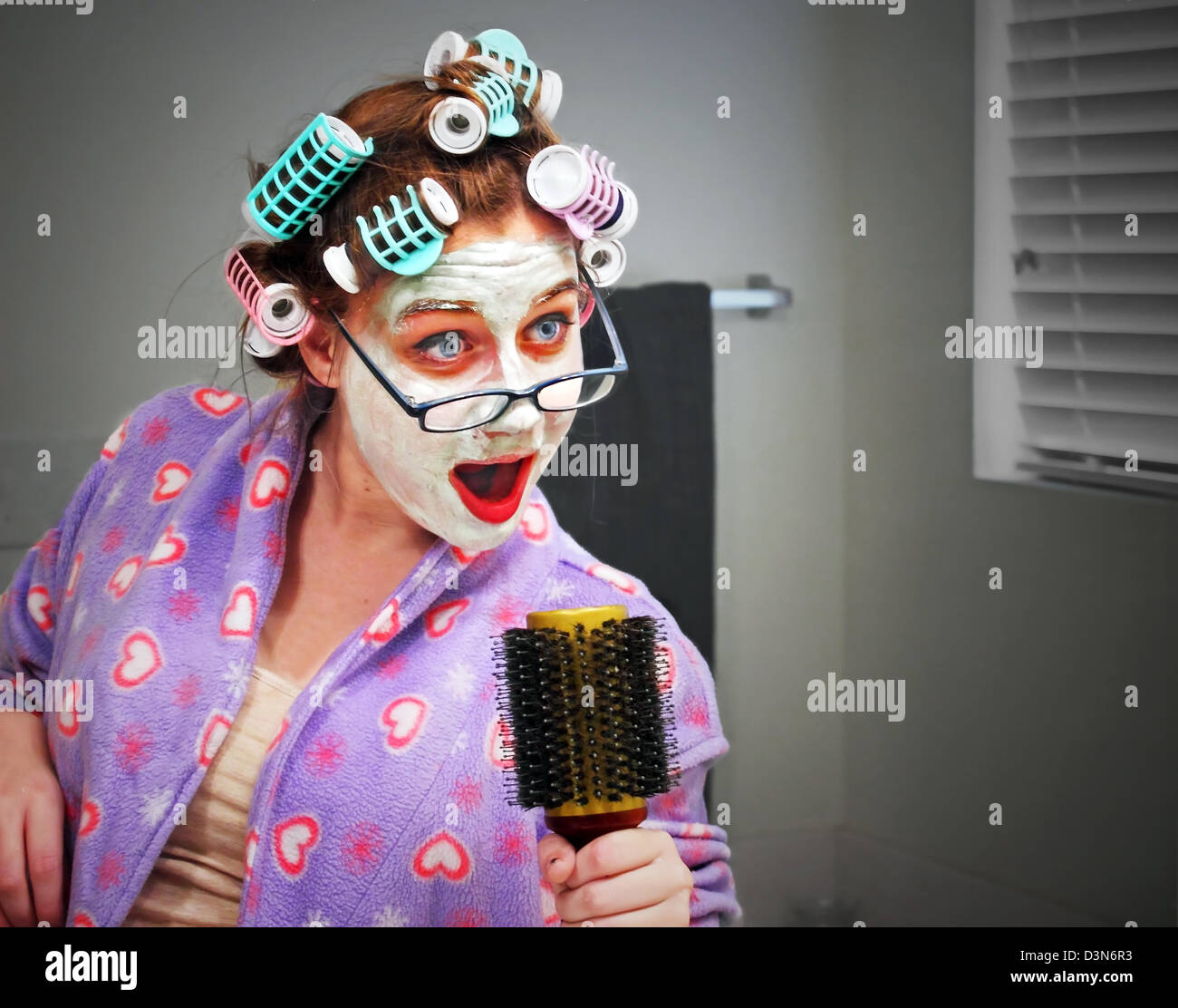 A young woman with glasses, curlers and a facial masque sings into a hair brush while preparing to head out on the town. Stock Photo