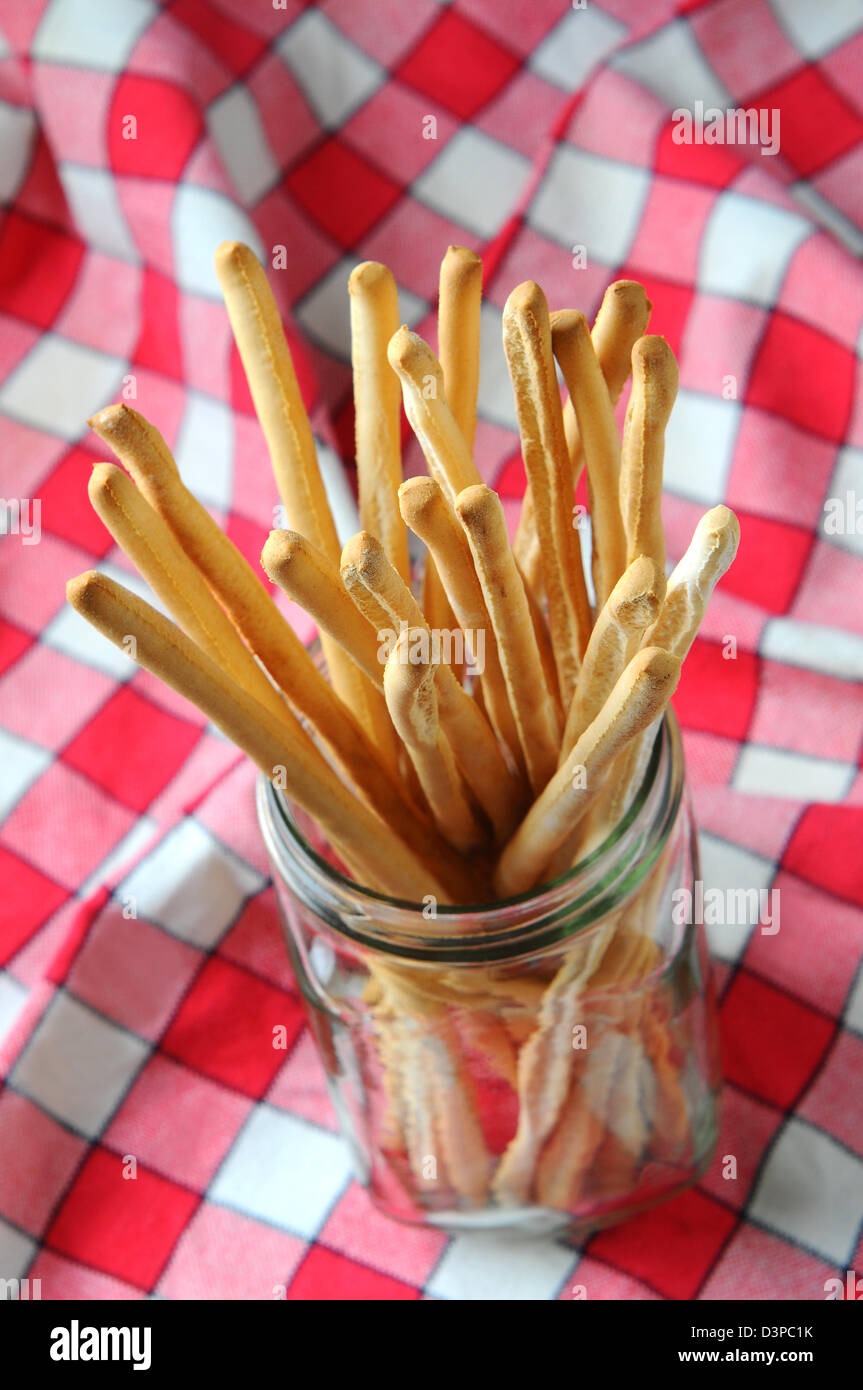 High angle closeup of bread sticks in a glass jar on a red and white checkered tablecloth. Vertical format. Stock Photo