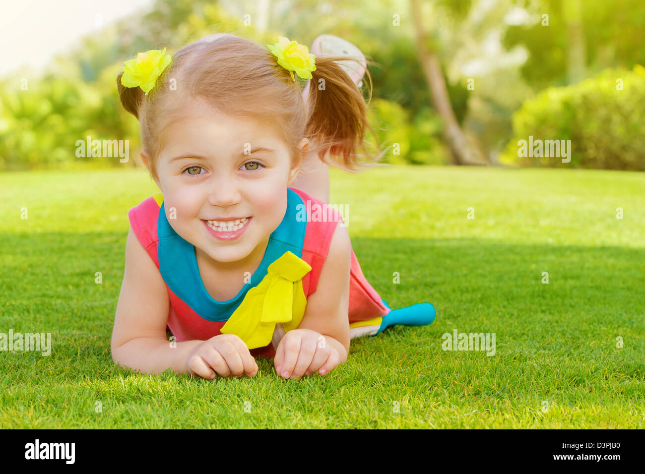 Picture of cute little girl lying down on green grass in park, cheerful child resting on the field on backyard, pretty kid Stock Photo