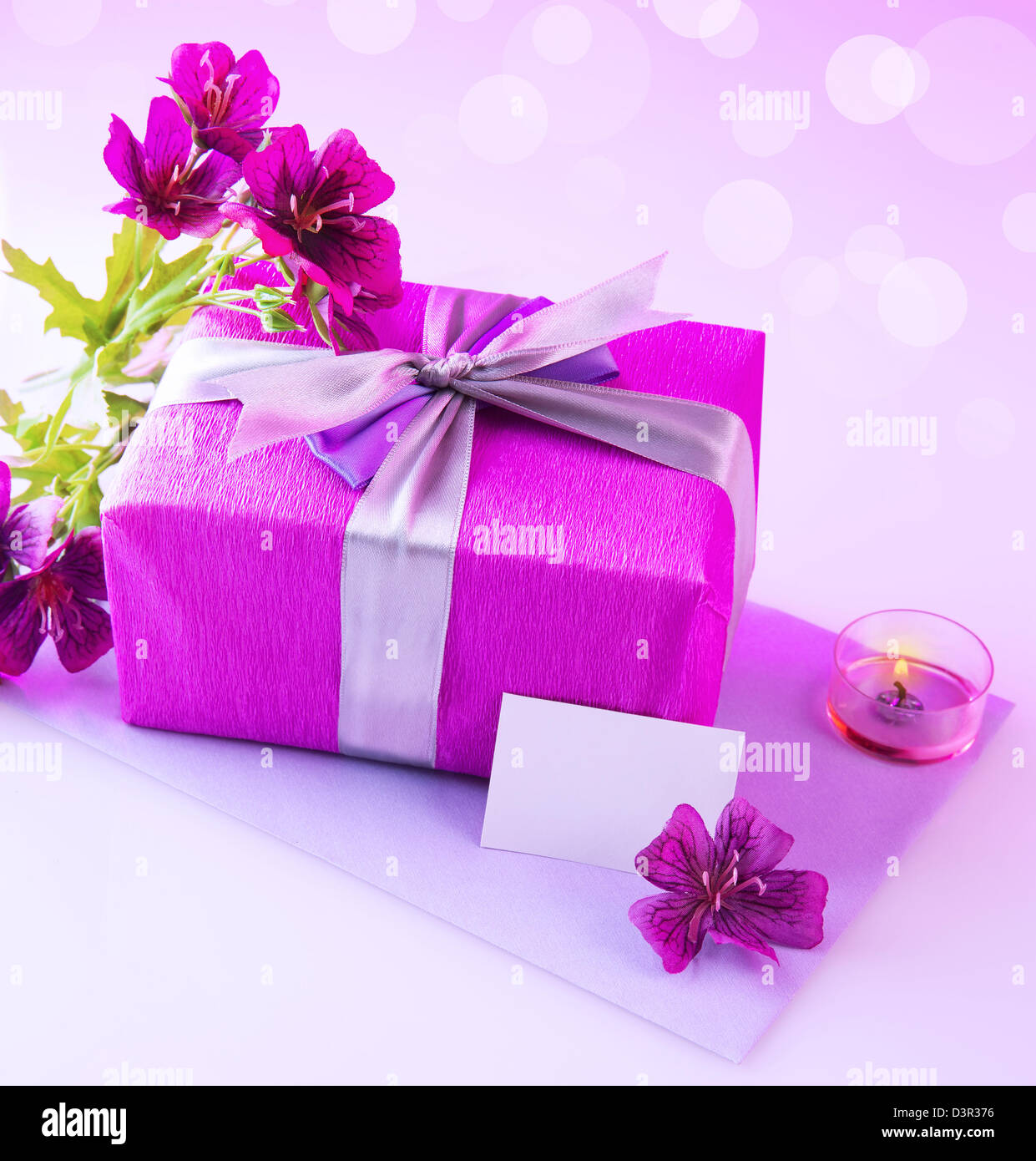 Photo of pink giftbox with silk bow, fresh purple flowers, candle and blank postcard isolated on blur background, festive still Stock Photo