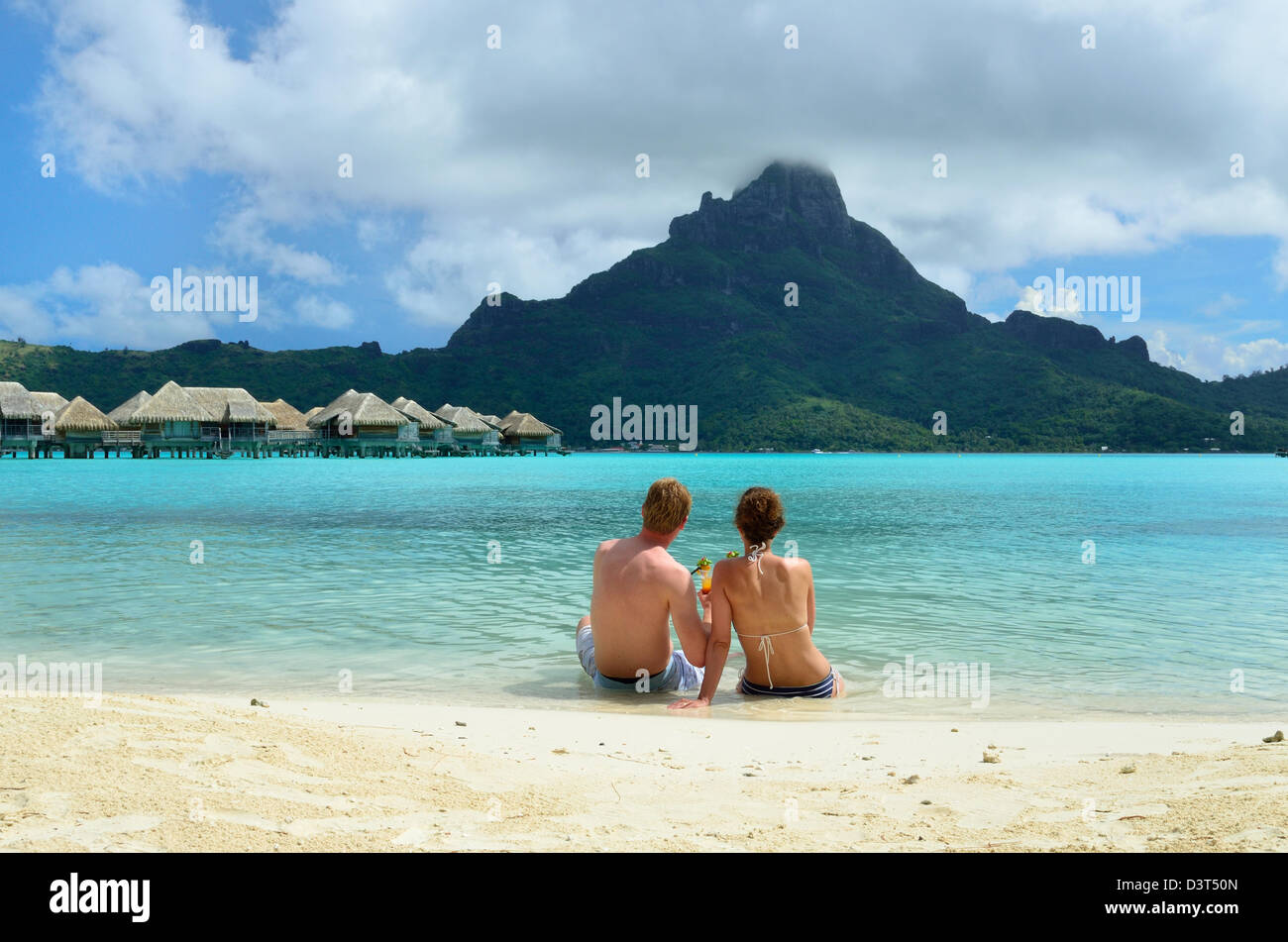 A honeymoon couple drinking a cocktail on the beach of a luxury vacation resort in the lagoon with a view on a tropical island. Stock Photo