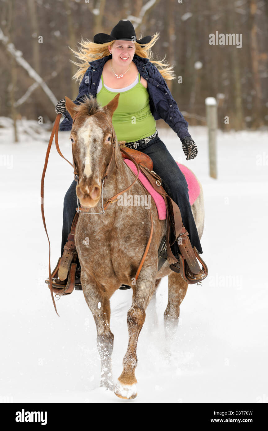 Woman riding Appaloosa horse in snowy field, blonde twentysomething wearing black cowboy hat, winter in Pennsylvania, PA, USA. Stock Photo