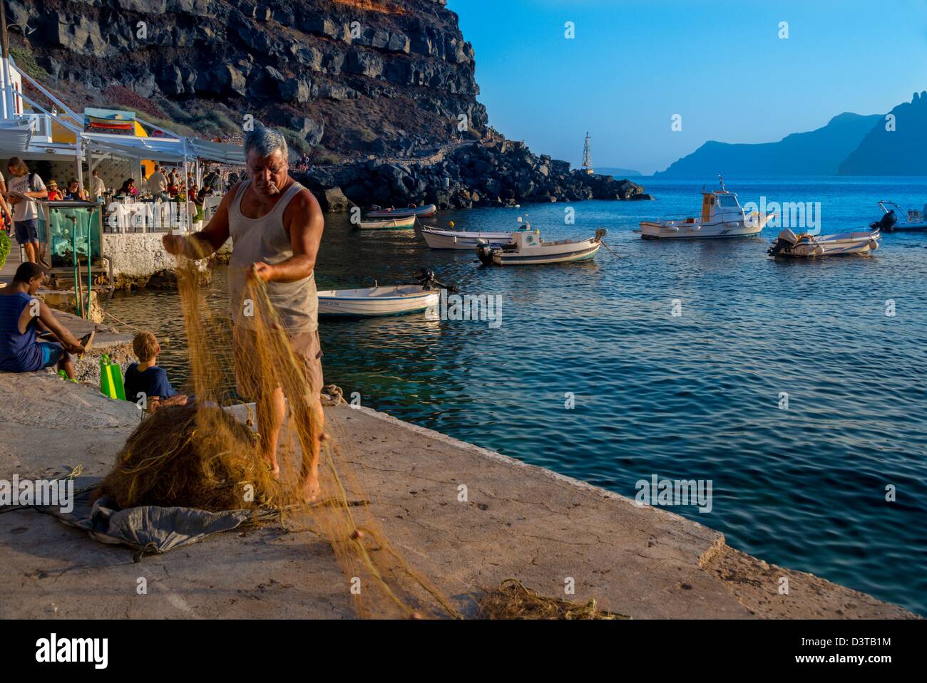 Fisherman prepares his net for fishing in Ammoudi, Santorini, Greece. Landscape view of port and fishing boats. Zorba the Greek Stock Photo