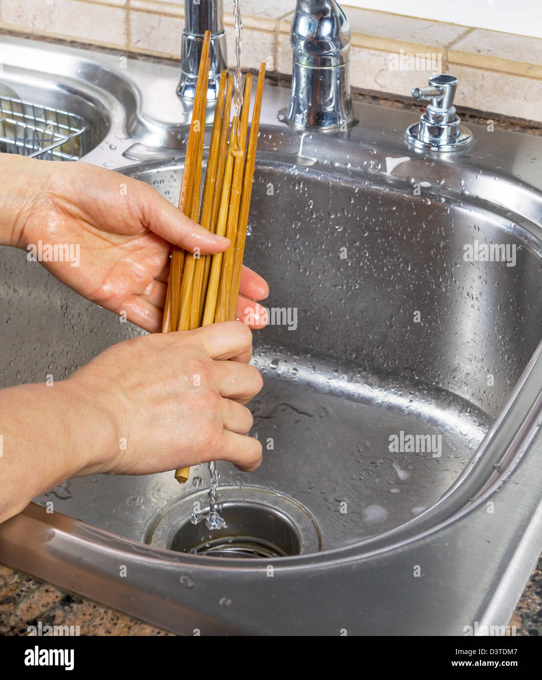 Vertical photo of female hands rinsing off bamboo chopsticks with running water from kitchen sink faucet in background Stock Photo