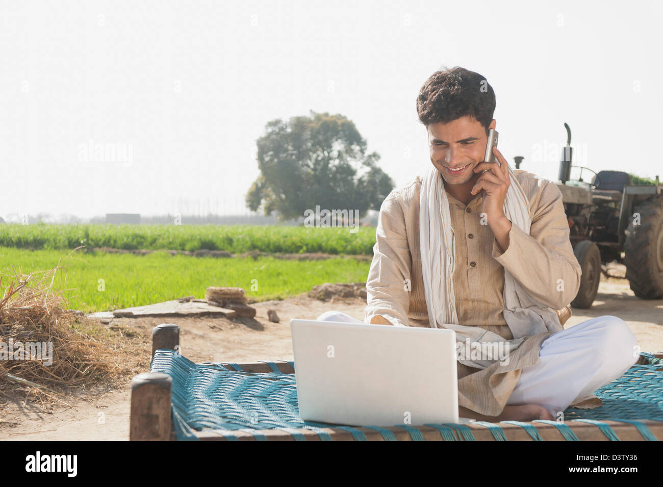 Farmer using a laptop and talking on a mobile phone in the field, Sonipat, Haryana, India Stock Photo