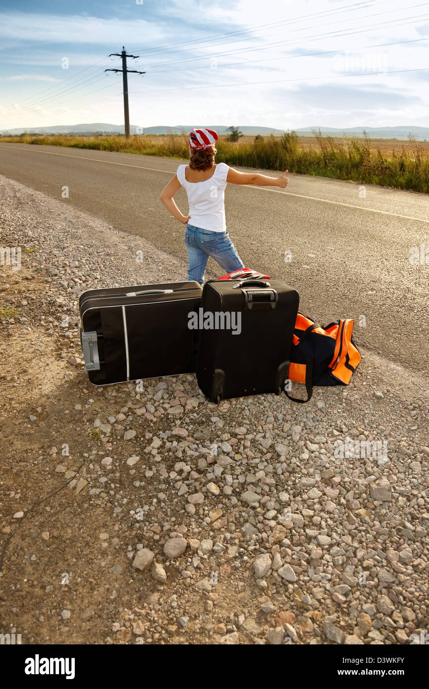 Young hitch-hiker girl standing on road side afternoon with suitcases Stock Photo