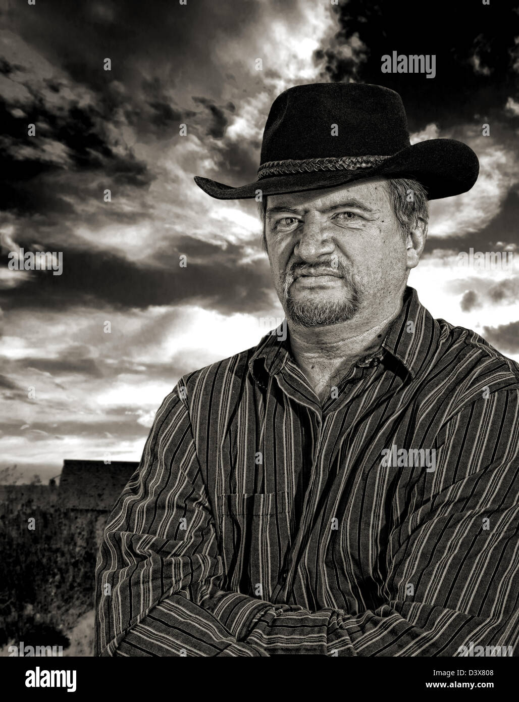 Rancher scowling with an old building with stormy sky in the background in monochrome with sepia toning Stock Photo