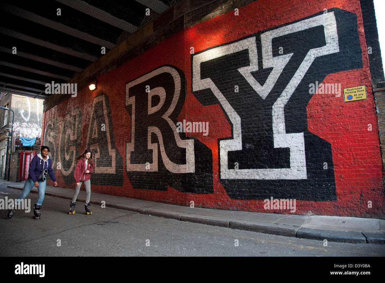 Urban girls rollerskate past the SCARY street art mural by Ben Eine in Shoreditch, East London. Stock Photo