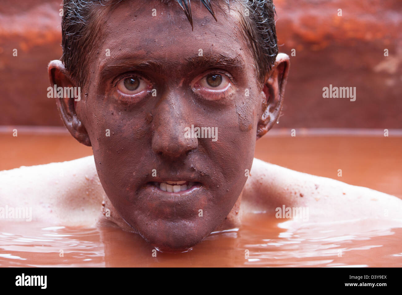 Close up of a man submerged in the red mud bath with his face completely covered in mud. Stock Photo