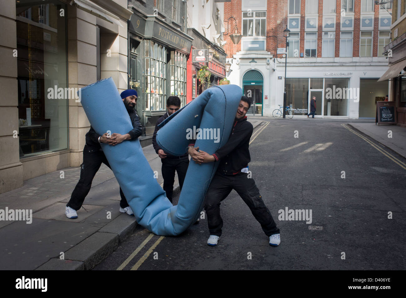 A Team Of Asian Workmen Manhandle Roll Industrial Carpet In Side Street And Destined For Nearby Shop Londons New Bond A Team Of Asian Workmen Manhandle Roll Industrial Carpet In Side Street And Destined For Nearby Shop Londons New Bond