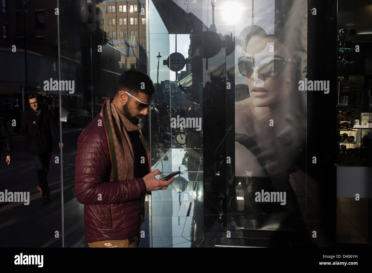 A man walks past a healthy poster girl for Burberry sunglasses they call  Eyewear, in a sunlit London street. With the model wearing the glasses  looking towards the young male, other Londoners