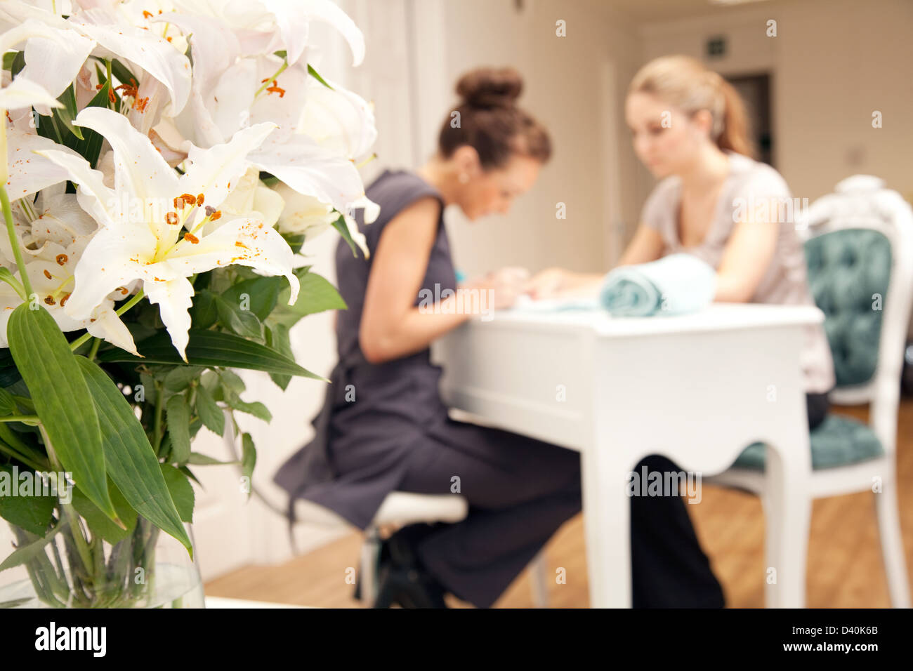 A young woman having her nails painted in a beauty salon. The subjects are in the background with a vase of flowers to the left Stock Photo