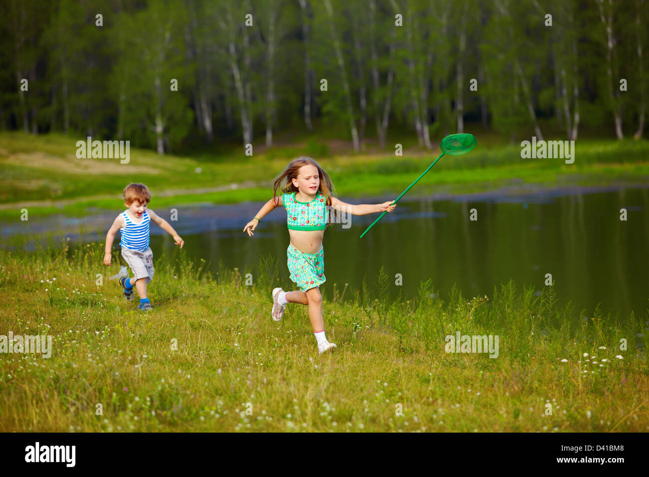 outdoor portrait of little kids running with butterfly net along the bank of the pond Stock Photo