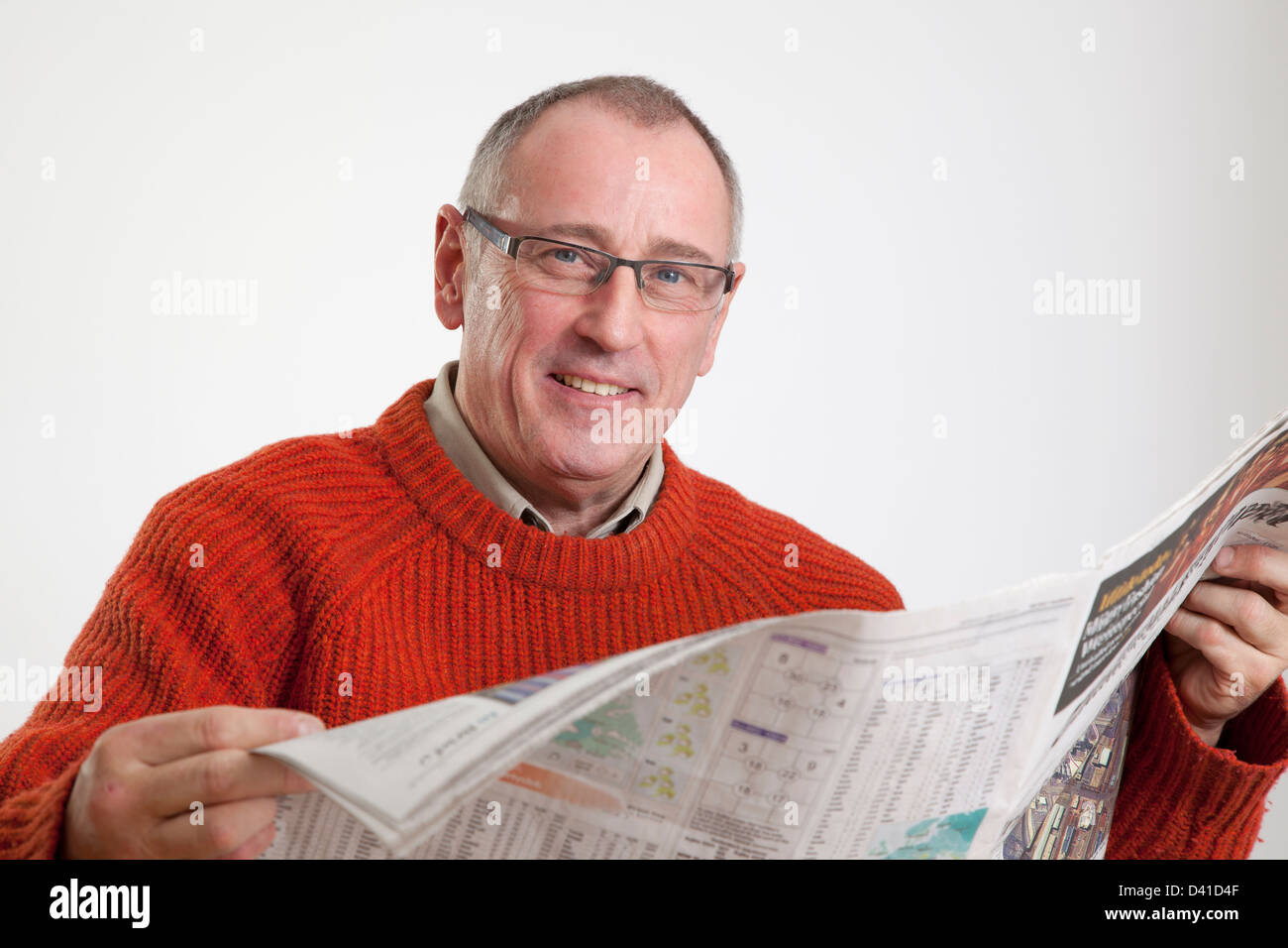 Mature man in 50s wearing sweater, reading a broadsheet newspaper, looking to camera smiling. Stock Photo