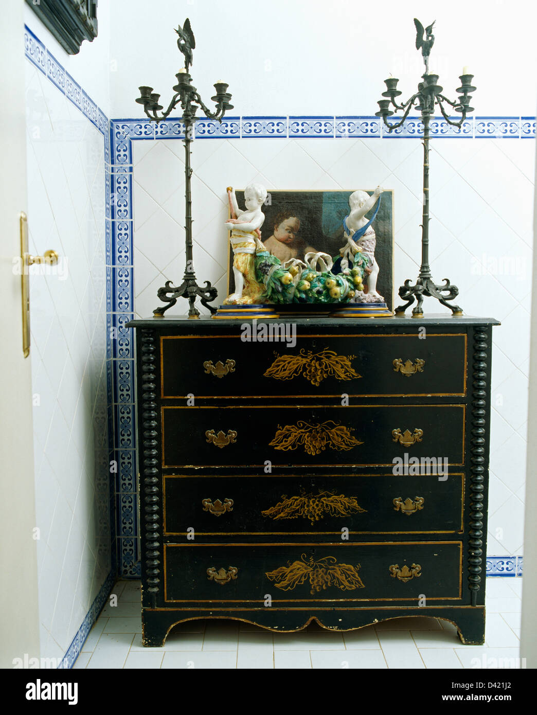 Ornate metal candlesticks and ceramic fruit on antique painted chest-of-drawers in bathroom with blue+white tiled border Stock Photo